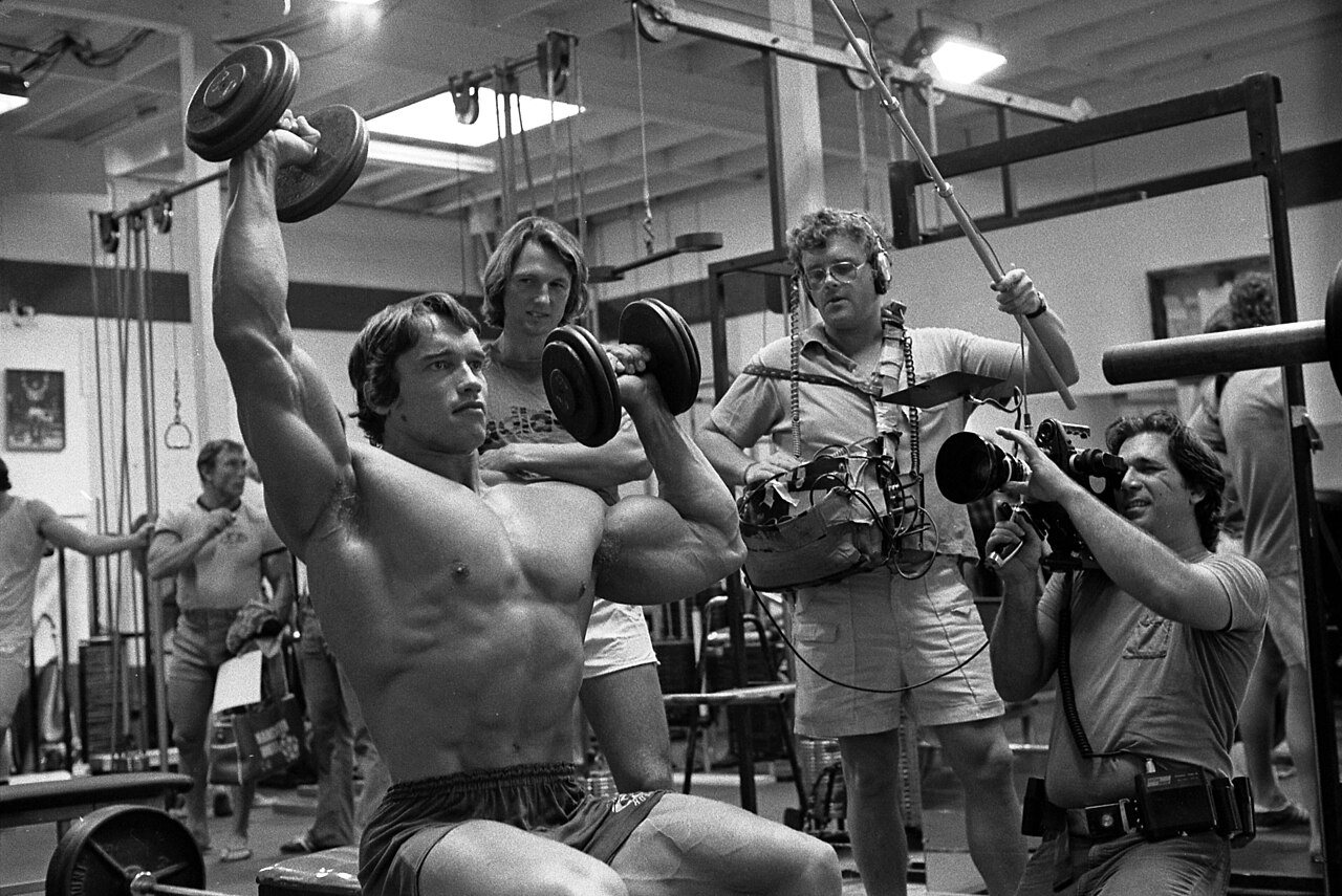 Arnold Schwarzenegger lifting dumbbells while a documentary crew films him at Gold's Gym in 1975